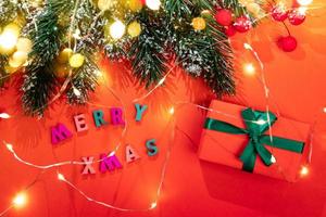 ramas de abeto, luces de navidad, nieve, bayas rojas, letras de colores feliz navidad, caja de regalo de bricolaje, luces de bokeh doradas en rojo. foto