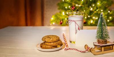 vaso de leche, galletas, nota vacía, adornos navideños en una mesa blanca contra un árbol de navidad con luces. foto