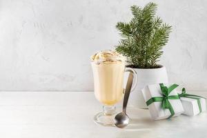 Glass of Christmas eggnog, spoon, small Xmas tree on flower pot, gift boxes on white. Copy space.