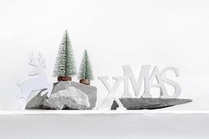 Xmas trendy modern composition. Christmas white decorations letters XMAS, deer, Xmas trees on stone stands on white. photo