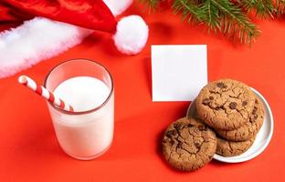 vaso de leche con pajita, galletas, nota vacía, ramas de abeto, gorro de papá noel en rojo. de cerca. foto