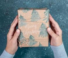 Women's hands holds gift box wrapped in festive wrapping paper with spruces and words Merry Christmas over turquoise. photo