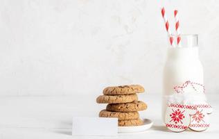 Bottle of milks, cookies, empty note, decorative patterned mitts on white table on white. Copy space photo