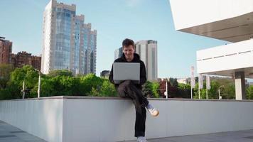 chico trabaja en una computadora portátil en un parque de la ciudad con el telón de fondo de la vegetación. hombre independiente usa cuaderno para trabajo al aire libre. concepto de trabajo remoto. camara lenta. video