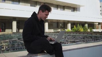 chico trabaja en una computadora portátil en un parque de la ciudad con el telón de fondo de la vegetación. hombre independiente usa cuaderno para trabajo al aire libre. concepto de trabajo remoto. camara lenta. video