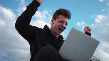 el trabajador independiente feliz se regocija del éxito usando una computadora portátil en el parque de la ciudad en un día soleado. el hombre alegre está encantado de ganar un juego en línea. victoria de las emociones. la cámara gira 360 grados. camara lenta. video