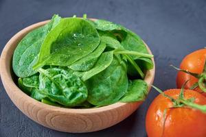 Fresh spinach leaves in wooden bowl and tomato on dark background. Organic food photo