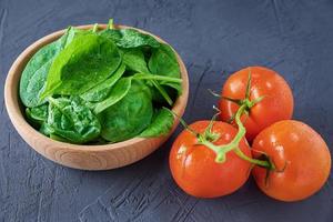 Fresh spinach leaves in wooden bowl and tomato on dark background. Organic food photo