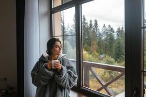 Woman holding mug, sitting on windowsill and looking at mountains photo