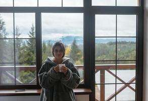 mujer joven de pie junto a la ventana sostiene una taza foto