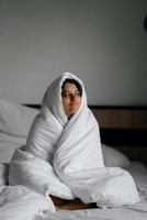 mujer perezosa envuelta en una manta suave sentada en una cama acogedora foto