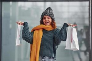 sonriendo y sintiéndome feliz. hermosa chica alegre con bufanda amarilla y ropa abrigada de pie en el interior con bolsas de compras en las manos foto