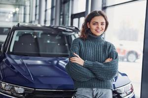 Cheerful young woman standing in front of modern new car indoors photo