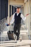 Male passenger in elegant formal clothes is in the airport hall with baggage, tickets and phone photo