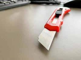 papelería en la mesa de la oficina. cuchillo de papelería con mango de plástico rojo. corte de papel, herramienta para el trabajo. cuchillo con una hoja afilada de metal foto