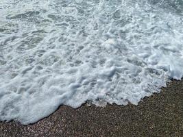 olas de agua en el mar y arena con pequeñas piedras multicolores naturales en la orilla del mar, pequeños guijarros en la playa. fondo, textura foto