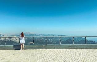 a girl with long hair in a white dress on a viewing platform on top of a mountain. hiking trip. girl watching the mountains from above photo