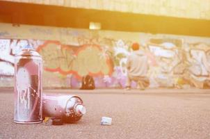 Several used spray cans with pink and white paint lie on the asphalt against the standing guy in front of a painted wall in colored graffiti drawings photo