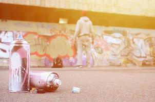 Several used spray cans with pink and white paint lie on the asphalt against the standing guy in front of a painted wall in colored graffiti drawings photo