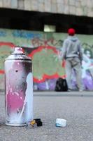 Used aerosol paint spray can with pink and white paint lie on the asphalt against the standing guy in front of a painted wall in graffiti drawings photo