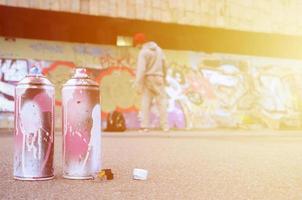 Several used spray cans with pink and white paint lie on the asphalt against the standing guy in front of a painted wall in colored graffiti drawings photo