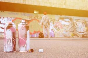 Several used spray cans with pink and white paint lie on the asphalt against the standing guy in front of a painted wall in colored graffiti drawings photo