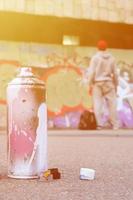 Used aerosol paint spray can with pink and white paint lie on the asphalt against the standing guy in front of a painted wall in graffiti drawings photo
