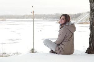 A young Caucasian girl in a brown coat is sitting near a cliff in the background of a horizon line between the sky and a frozen lake in winter time photo