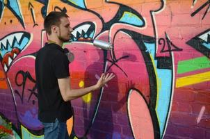 Young graffiti artist with gas mask on his neck throw his spray can against colorful pink graffiti on brick wall. Street art and contemporary painting process photo