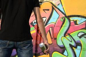 Young graffiti artist with backpack and gas mask on his neck paints colorful graffiti in pink tones on brick wall. Street art and contemporary painting process photo