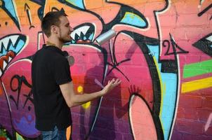 Young graffiti artist with gas mask on his neck throw his spray can against colorful pink graffiti on brick wall. Street art and contemporary painting process photo