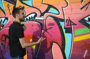 Young graffiti artist with gas mask on his neck throw his spray can against colorful pink graffiti on brick wall. Street art and contemporary painting process photo