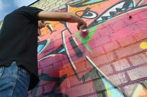 Young graffiti artist with backpack and gas mask on his neck paints colorful graffiti in pink tones on brick wall. Street art and contemporary painting process photo