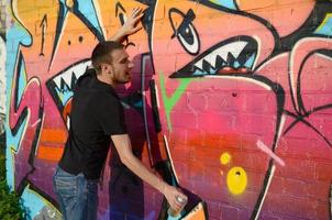 joven artista de graffiti caucásico en camiseta negra con lata de aerosol plateado cerca de graffiti colorido en tonos rosas en la pared de ladrillo. proceso de arte callejero y pintura contemporánea foto