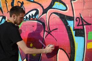 Young graffiti artist with backpack and gas mask on his neck paints colorful graffiti in pink tones on brick wall. Street art and contemporary painting process photo