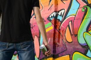 Young graffiti artist with backpack and gas mask on his neck paints colorful graffiti in pink tones on brick wall. Street art and contemporary painting process photo