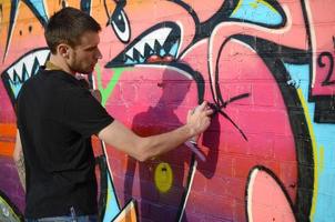 joven artista de graffiti con mochila y máscara de gas en el cuello pinta graffiti colorido en tonos rosas en la pared de ladrillo. proceso de arte callejero y pintura contemporánea foto