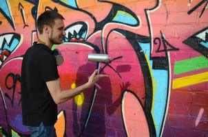 el joven artista de graffiti con máscara de gas en el cuello lanza su lata de aerosol contra el colorido graffiti rosa en la pared de ladrillo. proceso de arte callejero y pintura contemporánea foto