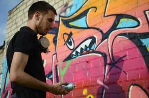 Young graffiti artist with backpack and gas mask on his neck paints colorful graffiti in pink tones on brick wall. Street art and contemporary painting process photo