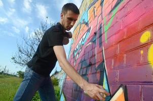 Young graffiti artist with backpack and gas mask on his neck paints colorful graffiti in pink tones on brick wall. Street art and contemporary painting process photo