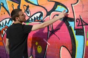 Young graffiti artist with backpack and gas mask on his neck paints colorful graffiti in pink tones on brick wall. Street art and contemporary painting process photo