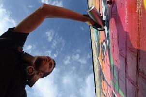 joven artista de graffiti con mochila y máscara de gas en el cuello pinta graffiti colorido en tonos rosas en la pared de ladrillo. proceso de arte callejero y pintura contemporánea foto