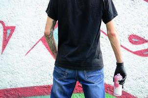 A young hooligan with a spray can stands against a concrete wall with graffiti paintings. Illegal vandalism concept. Street art photo