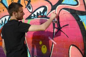 Young graffiti artist with backpack and gas mask on his neck paints colorful graffiti in pink tones on brick wall. Street art and contemporary painting process photo