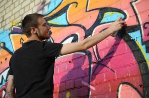 joven artista de graffiti con mochila y máscara de gas en el cuello pinta graffiti colorido en tonos rosas en la pared de ladrillo. proceso de arte callejero y pintura contemporánea foto