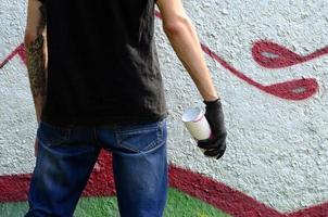 A young hooligan with a spray can stands against a concrete wall with graffiti paintings. Illegal vandalism concept. Street art photo