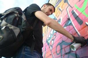 Young graffiti artist with backpack and gas mask on his neck paints colorful graffiti in pink tones on brick wall. Street art and contemporary painting process photo