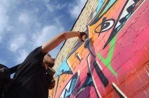 Young graffiti artist with backpack and gas mask on his neck paints colorful graffiti in pink tones on brick wall. Street art and contemporary painting process photo