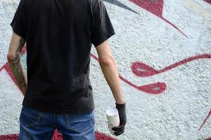 A young hooligan with a spray can stands against a concrete wall with graffiti paintings. Illegal vandalism concept. Street art photo