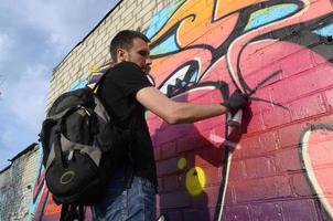 joven artista de graffiti con mochila y máscara de gas en el cuello pinta graffiti colorido en tonos rosas en la pared de ladrillo. proceso de arte callejero y pintura contemporánea foto